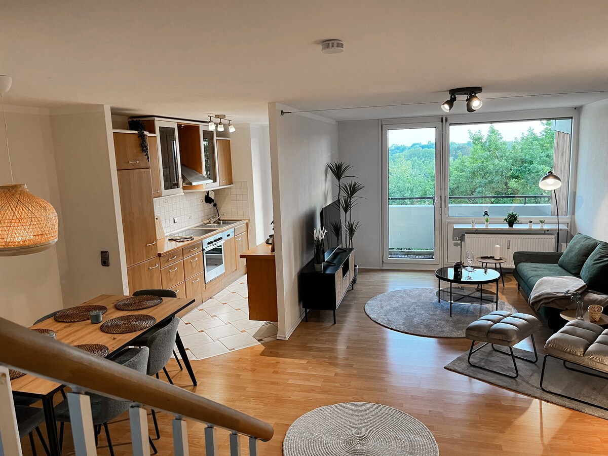 A bright living space showcases an open-plan design. The kitchen features wooden cabinetry and modern appliances, while the living area includes a green sofa and a round coffee table. A balcony is visible through large windows, framed by greenery outside. A dining table is set for four.