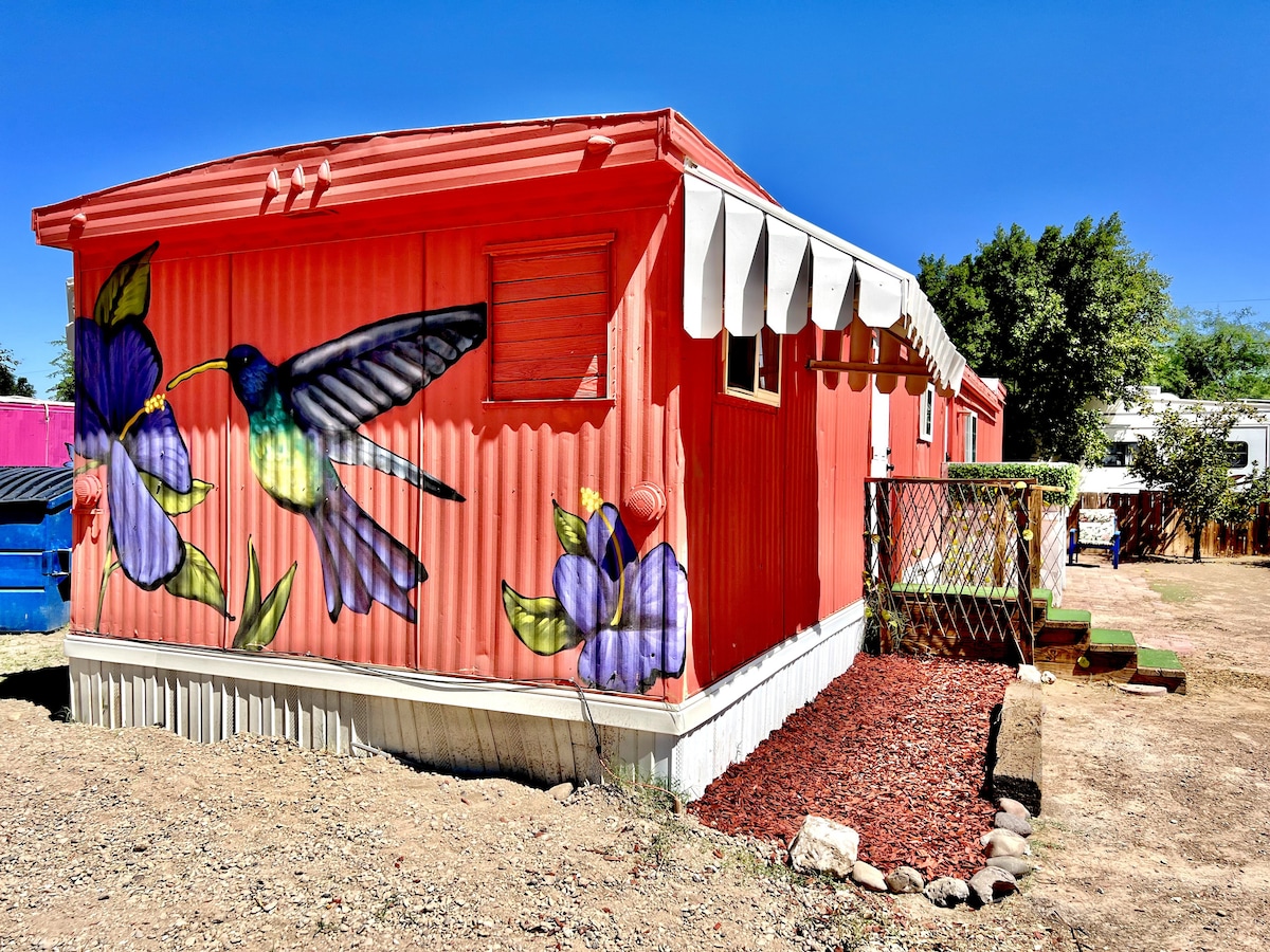 The exterior of a brightly colored trailer is adorned with a mural featuring a hummingbird and flowers. Sunlight highlights the vivid hues, while a well-maintained garden area with stone accents adds a touch of greenery beside the entryway.