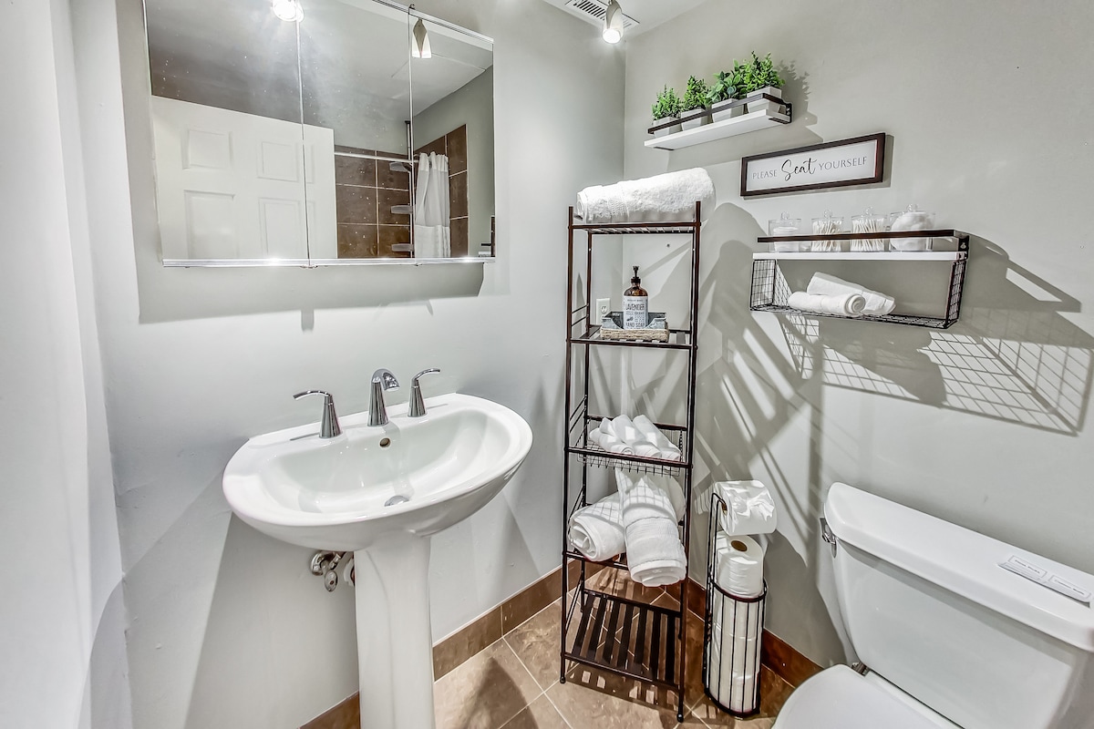 A functional bathroom is shown, featuring a mounted mirror above a modern sink with two faucets. A stylish metal shelving unit holds neatly arranged towels and toiletries, while a light-colored wall complements the warm tile flooring.