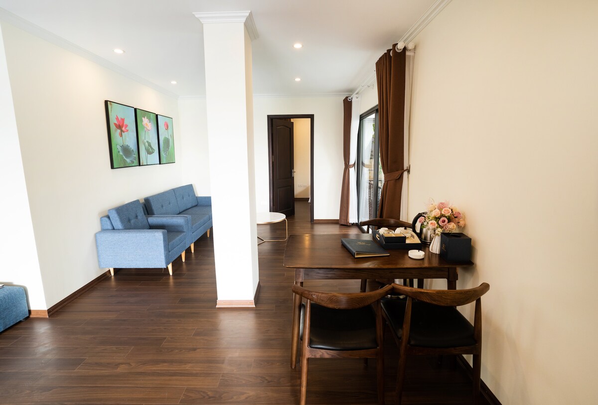 A spacious living area features a comfortable blue sofa and a wooden dining table with black chairs. Decorative flower art adorns the walls, while natural light filters through the partially closed curtains that frame a view of the outdoors.