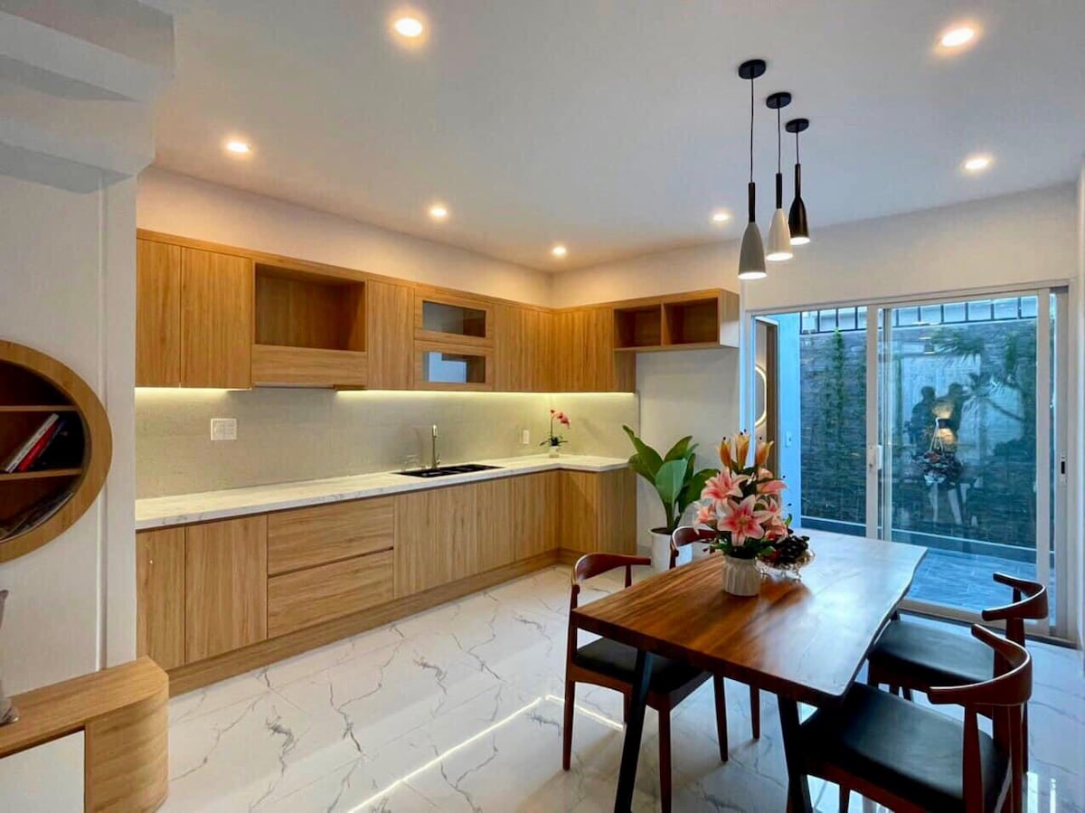 A modern kitchen showcases wooden cabinetry and a spacious countertop, with natural light illuminating the area. A dining table is present, paired with several chairs, while a view of the garden is offered through sliding glass doors.