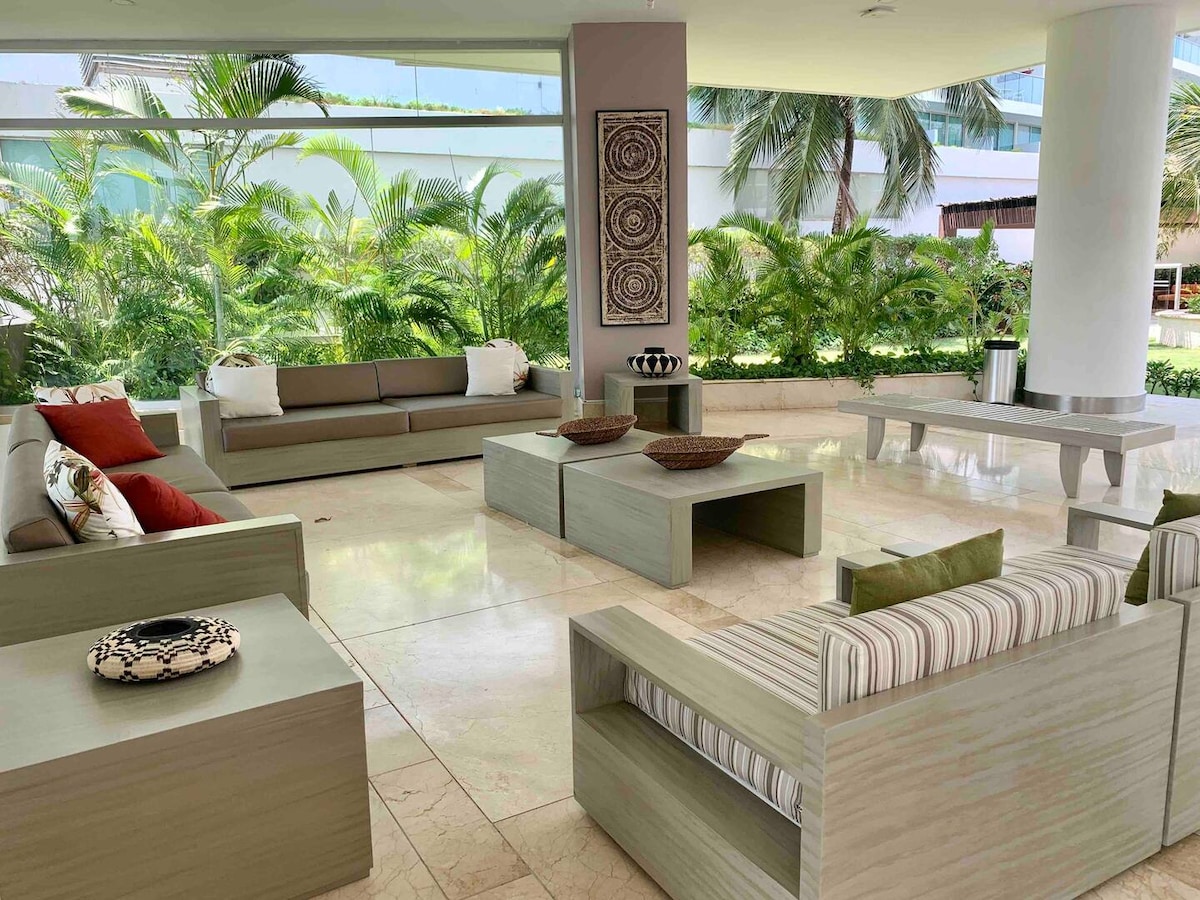 A spacious lounge area is presented with contemporary furniture, featuring low wooden coffee tables and comfortable seating options in neutral tones, accented by colorful cushions. Lush greenery is visible through expansive glass windows, enhancing the inviting atmosphere of the space.