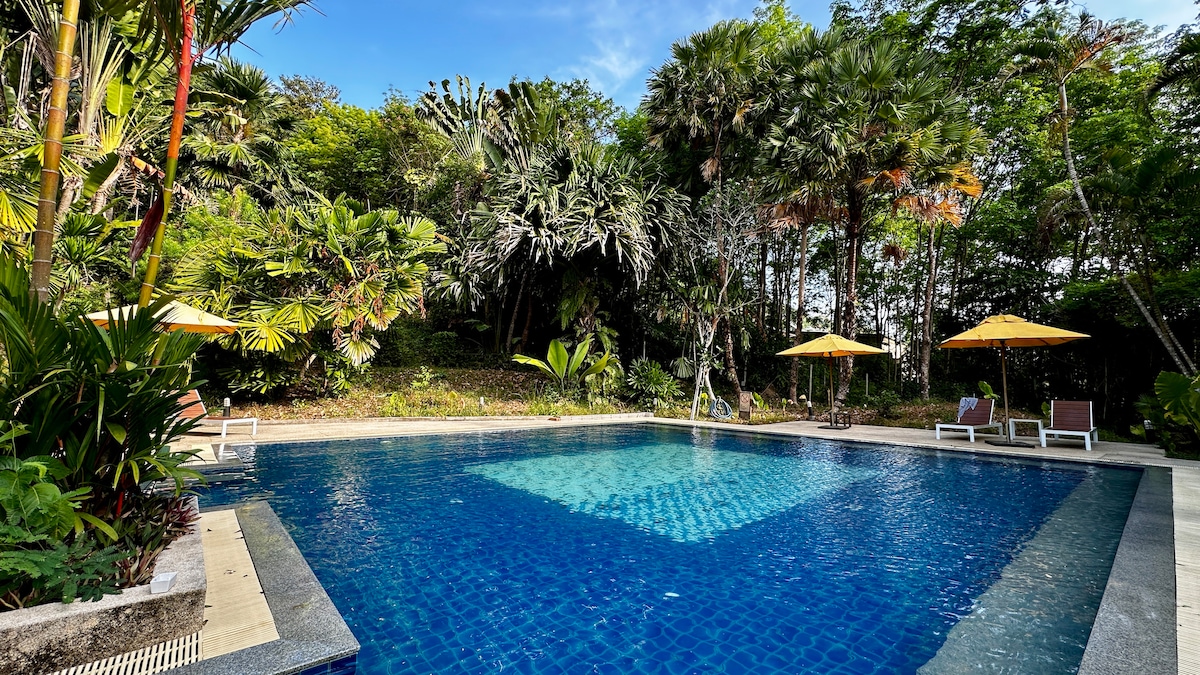 An outdoor swimming pool is surrounded by lush greenery and tropical plants. Sun loungers and umbrellas are positioned around the pool area, inviting relaxation. The clear water of the pool reflects the bright blue sky and the vibrant foliage, creating a serene escape.