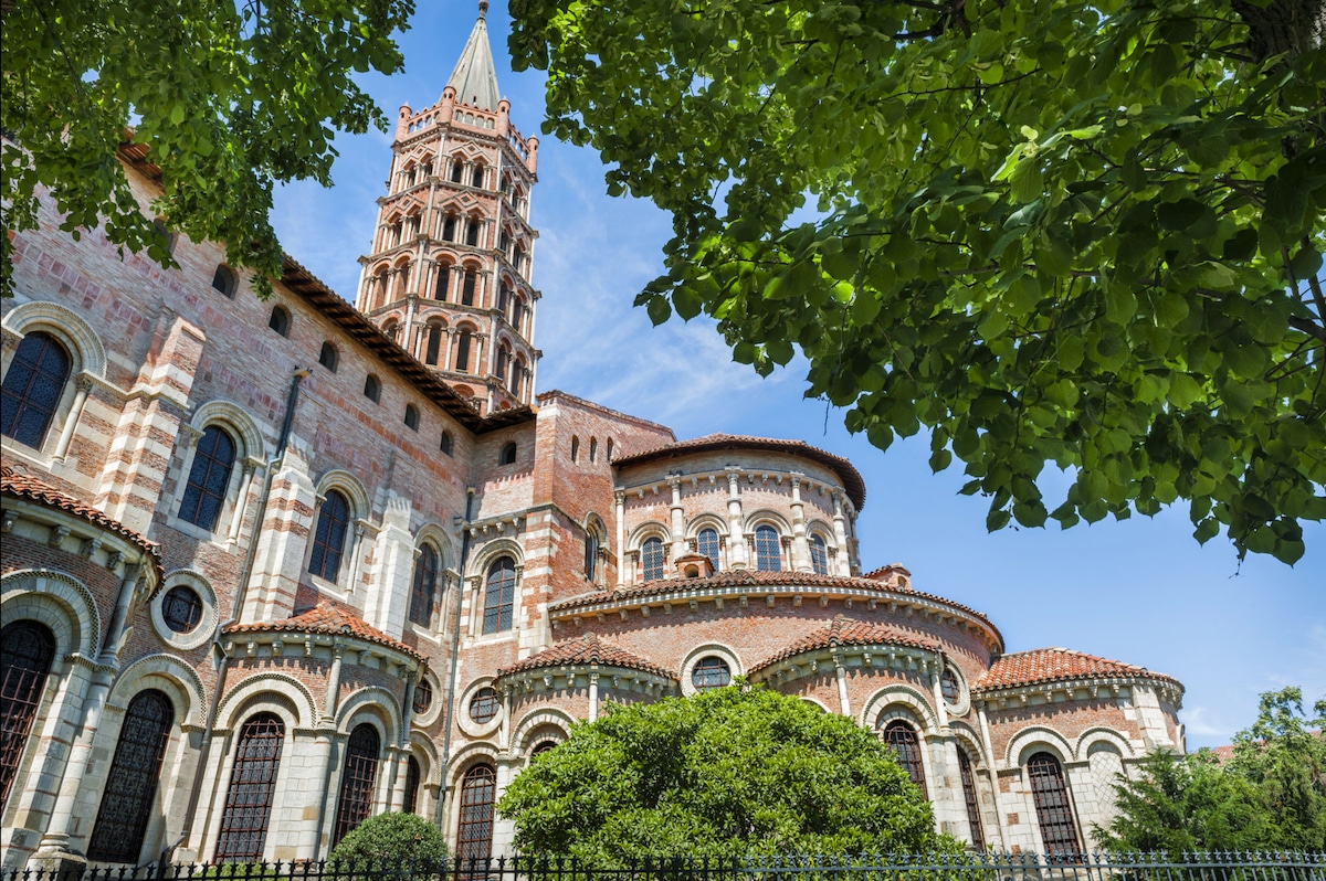 Historic architecture is showcased through a tall tower and intricately designed stonework. The building is framed by lush greenery, enhancing its grandeur. Bright blue skies provide a clear backdrop, highlighting the rich colors of the structure and its elaborate details.