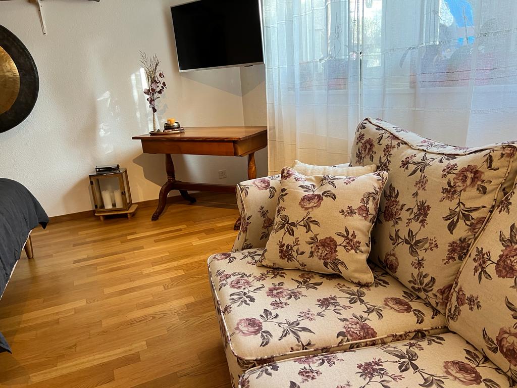 A floral-patterned sofa with plush cushions is positioned against a light wall. A wooden table with an elegant design sits nearby, complemented by a wall-mounted television. Soft natural light filters through sheer curtains, enhancing the warm wooden flooring.