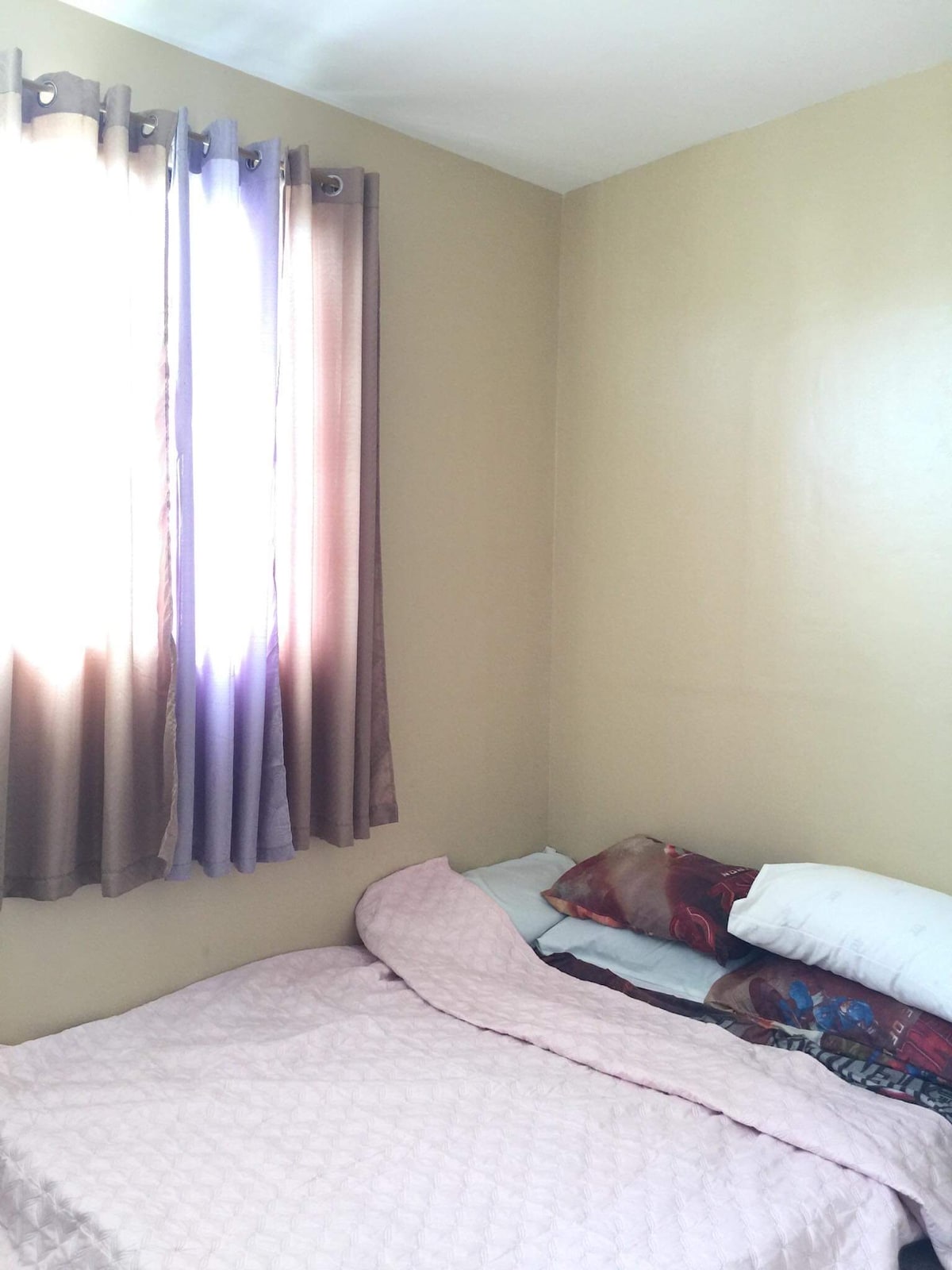 A small, cozy sleeping area features a soft pink quilt spread over a spacious bed. Several brightly patterned pillows are piled against the wall. Natural light filters through a window, covered by flowy, dark and light curtains.