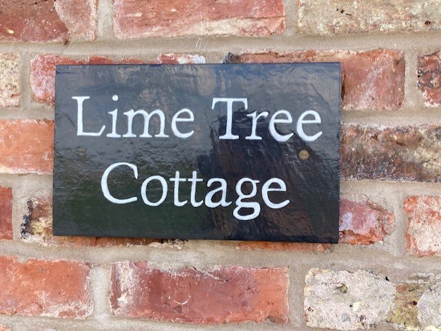 A sign reading 'Lime Tree Cottage' is affixed to a rustic brick wall. The sign features a dark background with white lettering, providing a clear indication of the cottage's name against the textured, earthy tones of the surrounding bricks.
