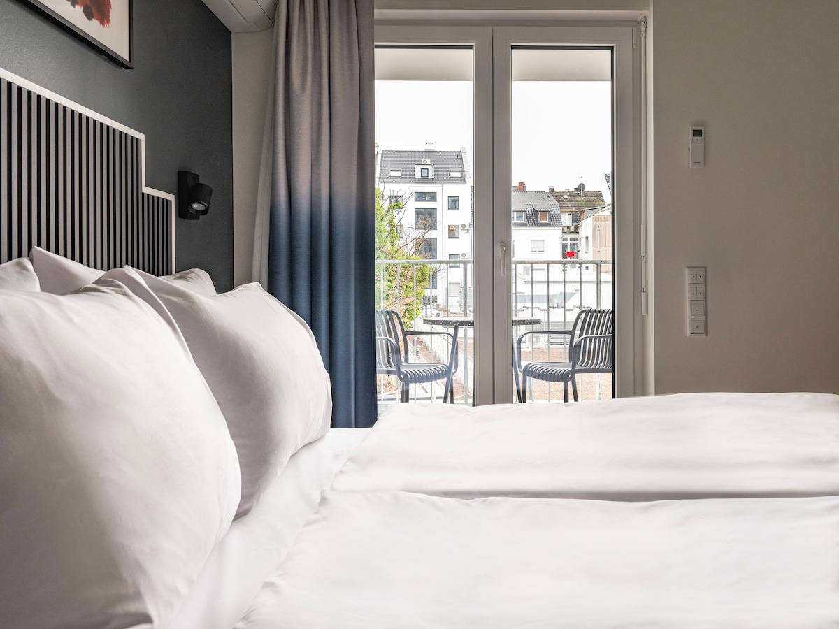 A spacious sleeping area features a double bed with white linens, complemented by soft pillows. Floor-to-ceiling glass doors open to a balcony, offering views of the outdoor space. Contemporary decor is seen in the muted color palette and wall detailing.