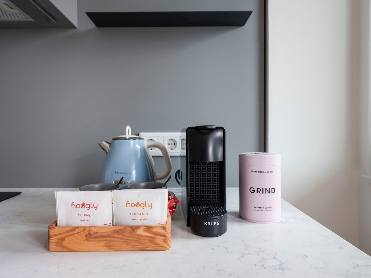 A modern kitchenette is showcased with a light marble countertop. A kettle in soft blue, a black coffee maker, and a pink coffee container are displayed. A wooden tray holds tea selections, contributing to the overall functionality of the space.