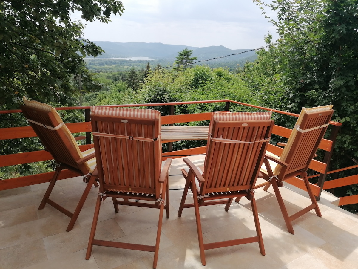 A balcony is furnished with four wooden lounge chairs and a small table, surrounded by a railing made of wooden slats. Lush greenery is visible in the background, along with a scenic view of distant hills and valleys under a cloudy sky.