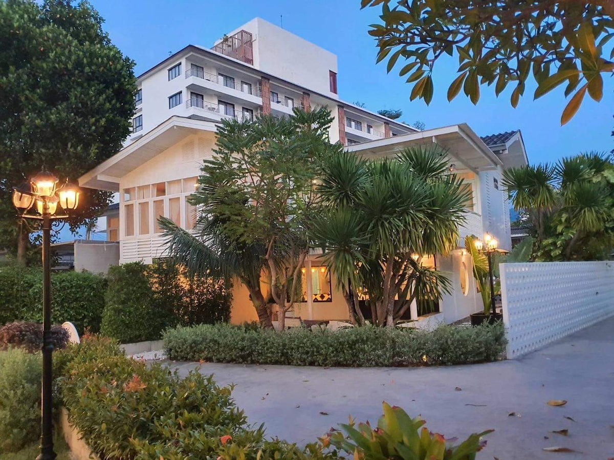 A spacious home is surrounded by lush greenery and tropical plants. Soft lighting illuminates the exterior, creating a welcoming ambiance. A modern building stands in the background, contrasting with the home's architectural design, while a well-maintained garden enhances the overall setting.