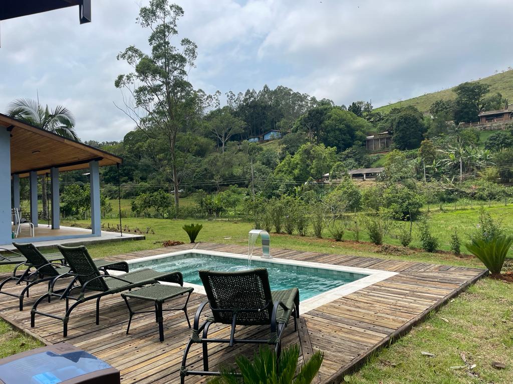 A serene outdoor area features a rectangular pool surrounded by wooden decking. Several lounge chairs are positioned nearby, providing space for relaxation. Lush greenery and gentle hills create a tranquil backdrop, enhancing the peaceful setting for family gatherings.
