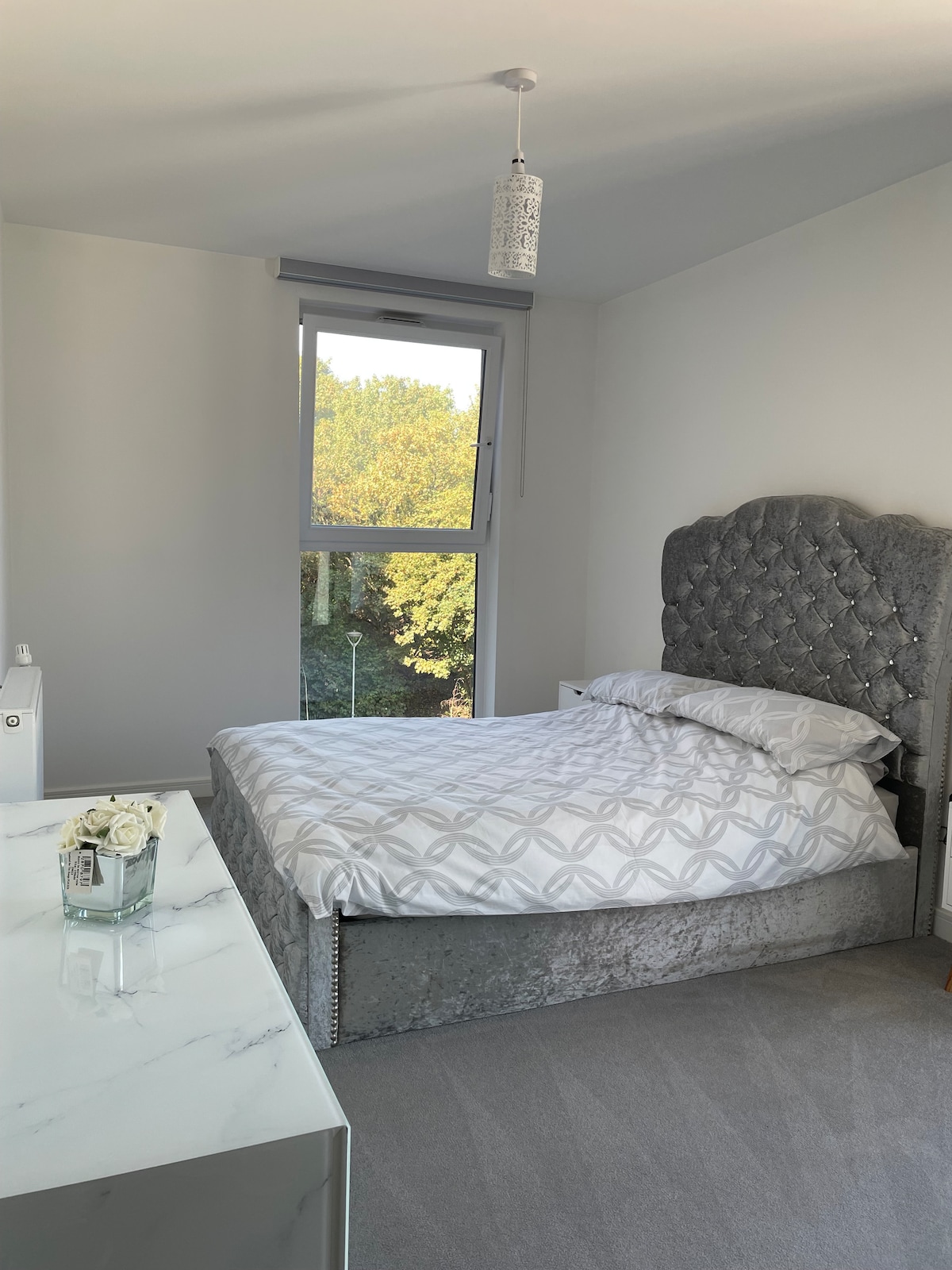 A serene bedroom features a spacious bed with a decorative grey headboard. Soft, patterned bedding is arranged neatly. Natural light enters through a large window overlooking lush greenery. A minimalist white dresser reflects a clean, modern aesthetic.