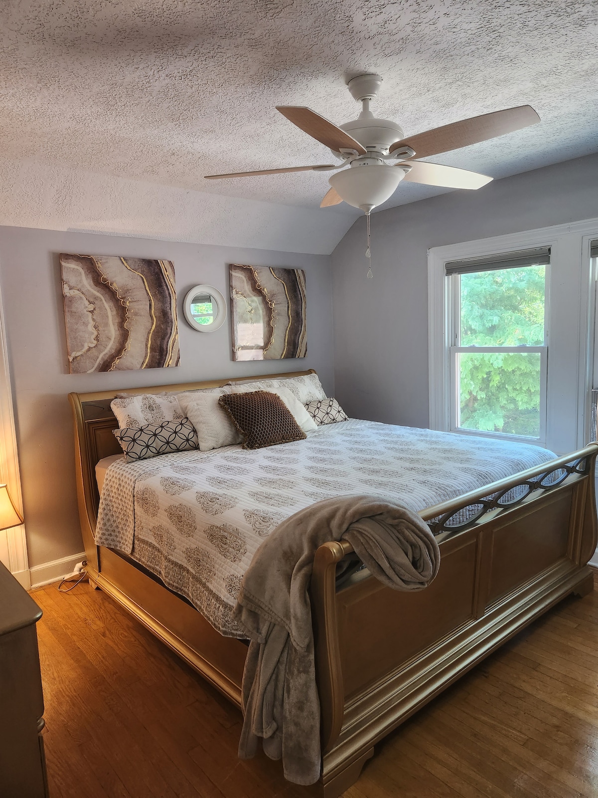 A spacious bedroom features a large bed with a decorative gray and white coverlet. Two abstract art pieces adorn the walls, and a bedside lamp provides soft lighting. Windows allow natural light and glimpses of greenery outside.