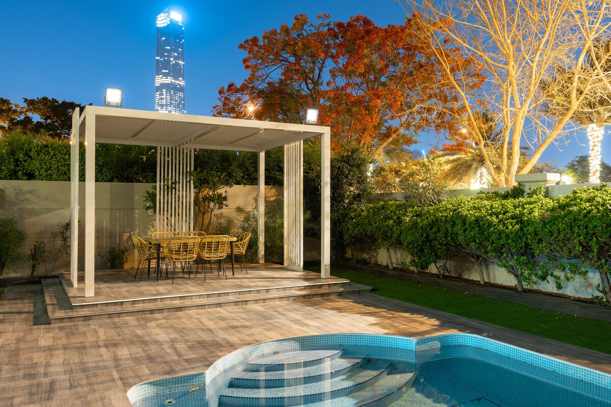 An outdoor dining area is featured under a modern pavilion, with a glass-topped table surrounded by stylish yellow chairs. A private pool glistens in the foreground, while lush greenery and city skyscrapers frame the serene setting, illuminated by soft evening lights.