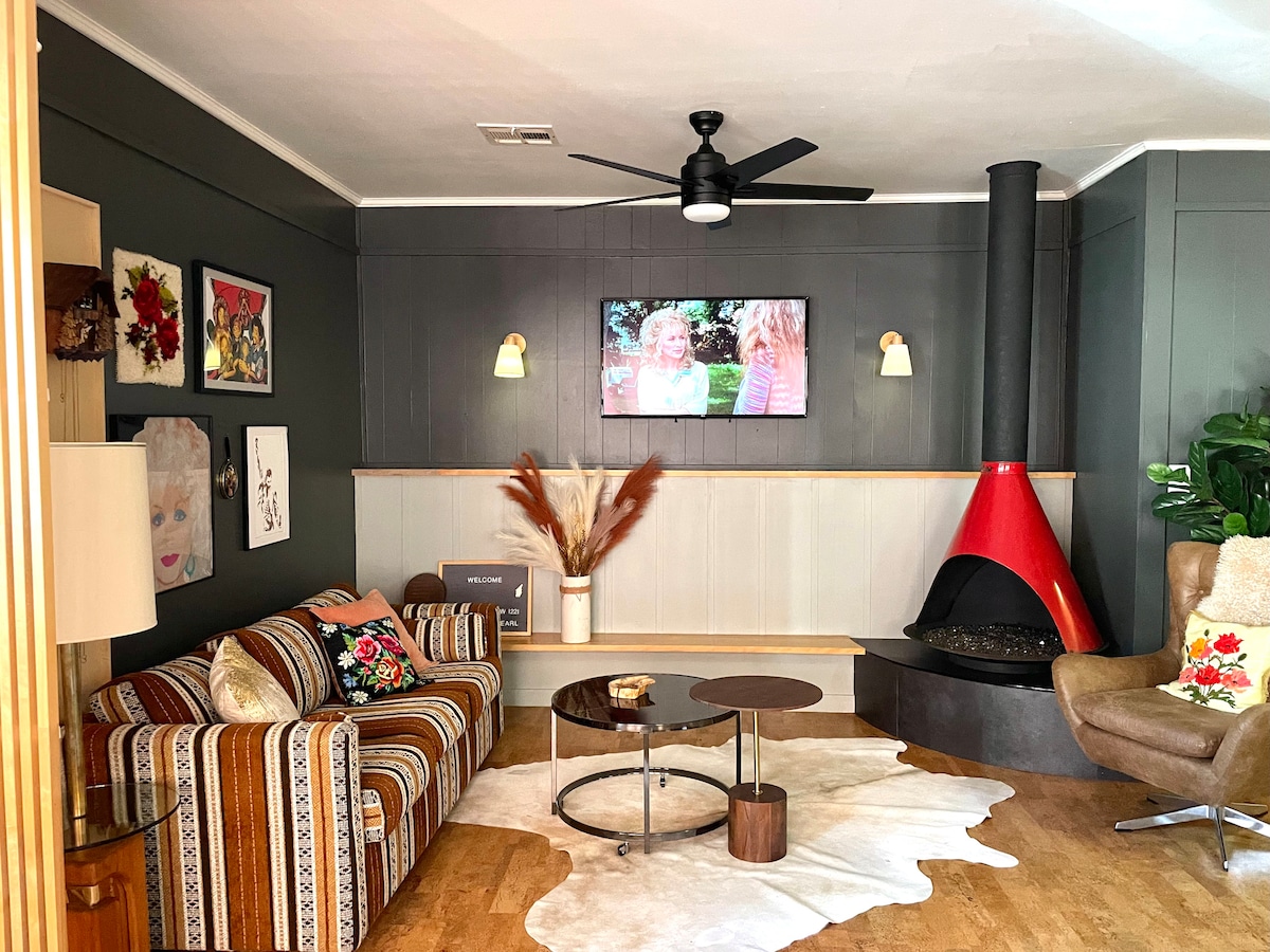 A living area features a striped sofa adorned with decorative pillows, complemented by a rustic coffee table. A red cone fireplace adds a bold touch, while framed art and plants enhance the ambiance. A ceiling fan and wall-mounted TV are visible, contributing to a cozy atmosphere.