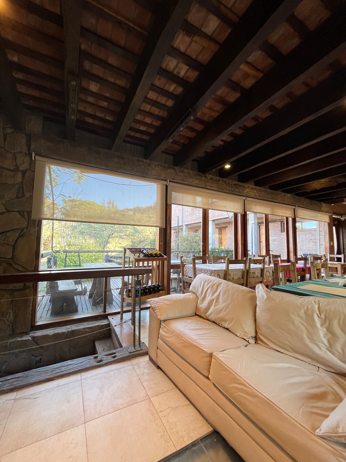 A spacious seating area features a light-colored sofa, providing comfort inside a rustic space. Large windows allow natural light to fill the room, while offering views of the outside greenery. Wooden beams and stone accents contribute to the cozy ambiance.