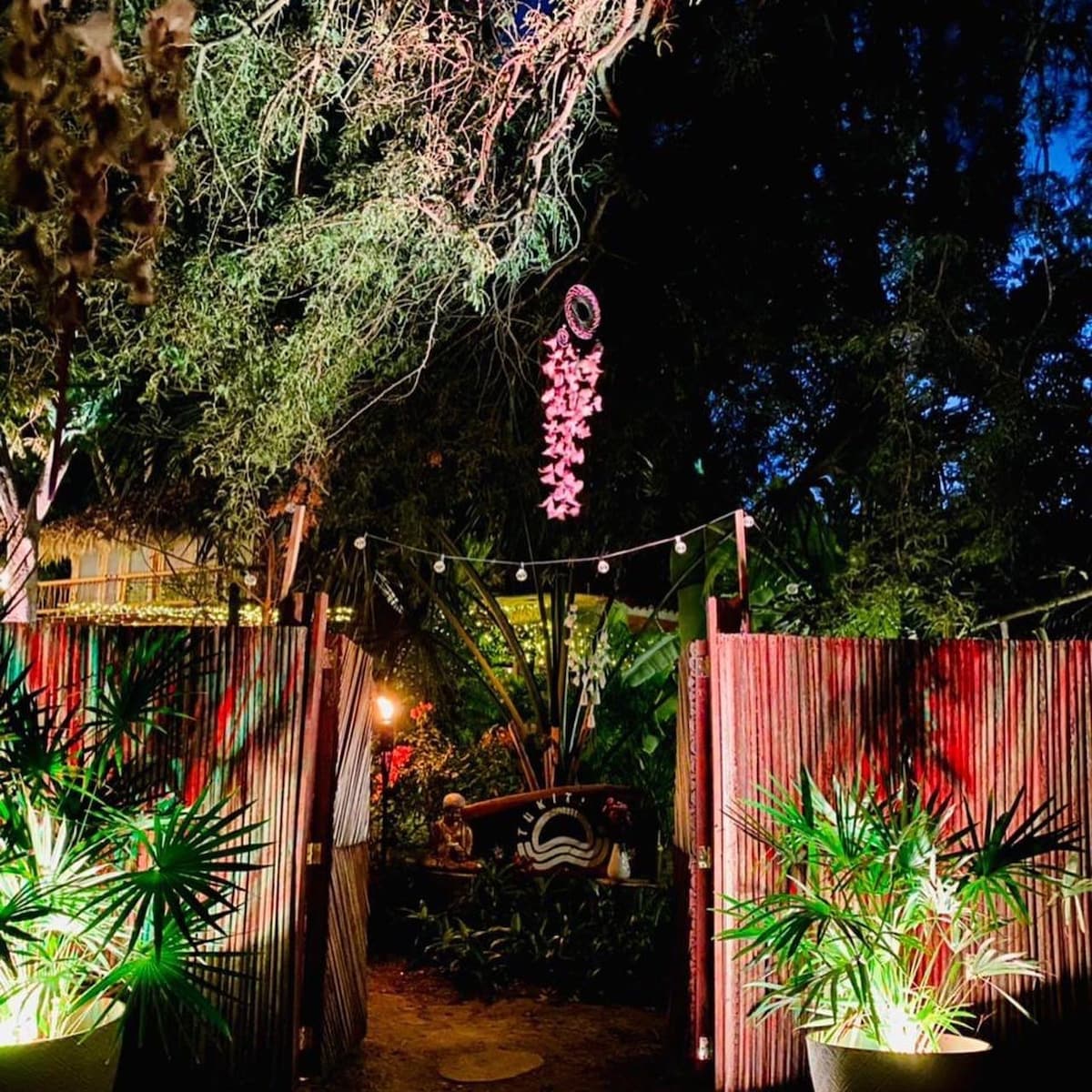 A charming entrance is framed by bamboo fencing and lush greenery, illuminated by soft lighting. A decorative mobile hangs above a pathway, guiding guests through a tropical setting. The inviting atmosphere is complemented by the surrounding plants and warm tones.