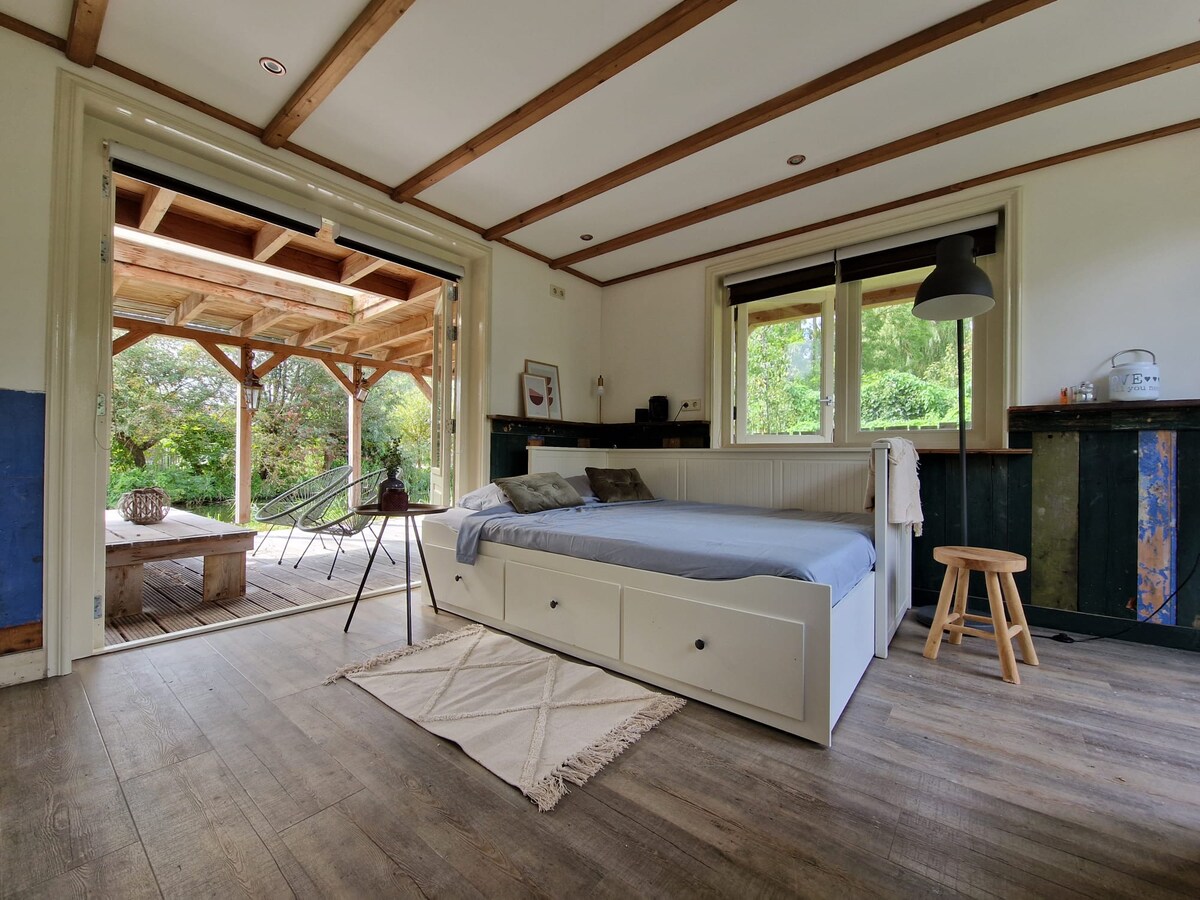 A studio space features a spacious bed with storage drawers underneath, complemented by a light rug on wooden flooring. Large windows provide views of the outdoor area, and a nearby light fixture hangs above. A small table and chairs are visible, enhancing functionality.