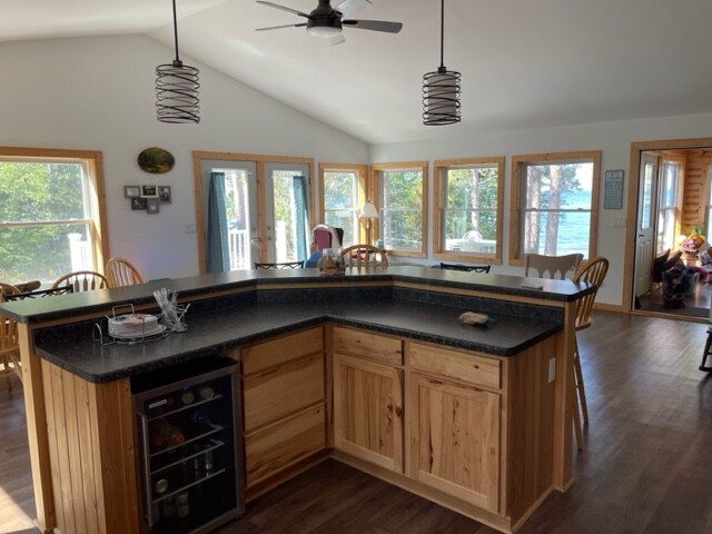 A bright kitchen area features wood cabinetry and a spacious island with a dark countertop. Large windows provide natural light and views of the outdoors. A dining table with chairs is visible, along with a small seating area in the background.