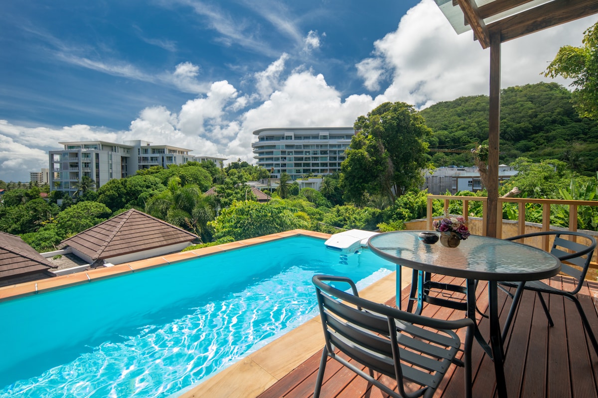 An inviting outdoor deck is shown, featuring a round dining table with four chairs. A vibrant pool captures sunlight, reflecting blue tones. Lush greenery and buildings are visible in the background under a bright sky, enhancing the serene atmosphere.