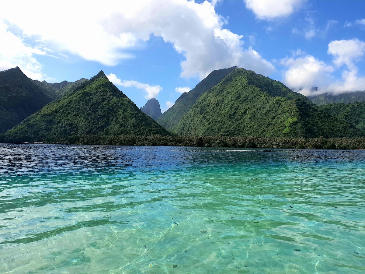 The image captures the serene lagoon with crystal-clear turquoise waters, reflecting the surrounding lush green mountains. Gentle waves ripple across the surface, while the expansive sky above features scattered clouds, enhancing the tranquil atmosphere of this natural landscape.