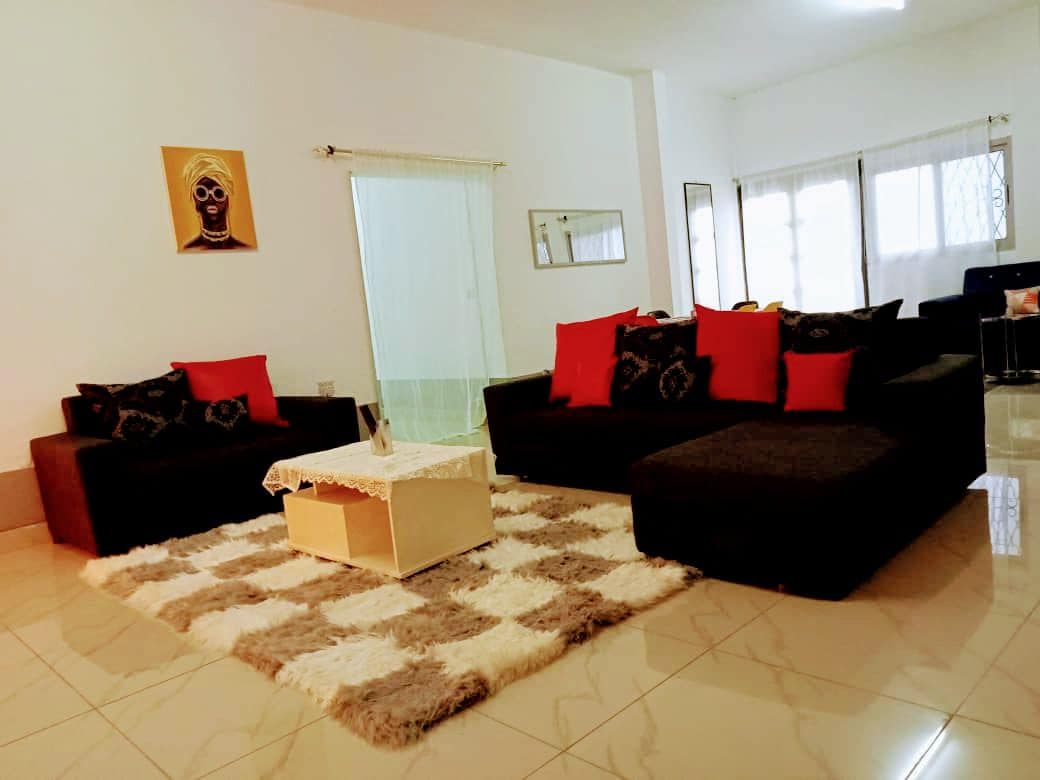 A spacious living area features two dark sofas adorned with red and black cushions. A coffee table sits atop a light, textured rug. Natural light filters through large windows, illuminating the room's neutral tones and contemporary decor.