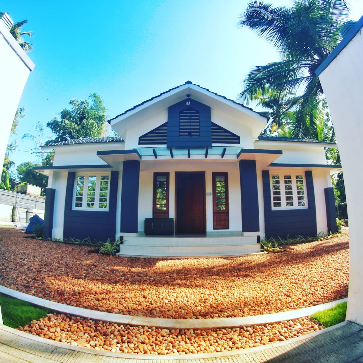 A charming villa is presented with a modern exterior, featuring a gabled roof and front porch. The entrance is framed by double doors, while multiple windows enhance natural light. A landscaped area of gravel and greenery surrounds the home, with tall palm trees in the background.