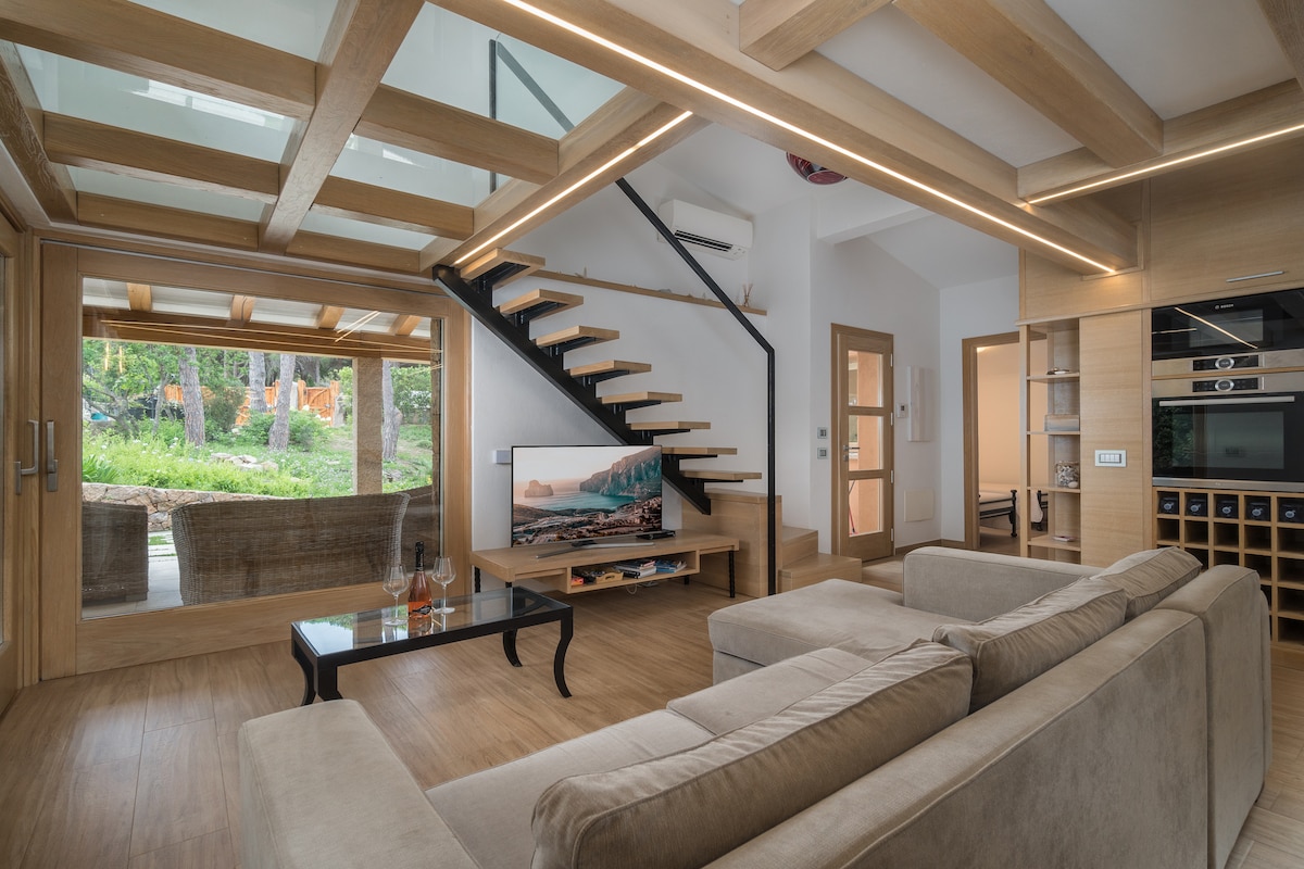 A modern living area features a comfortable sofa facing a low coffee table and an entertainment unit with a television. Natural light filters through large windows, showcasing a view of the outdoor space. A staircase with wooden steps leads to the upper level.