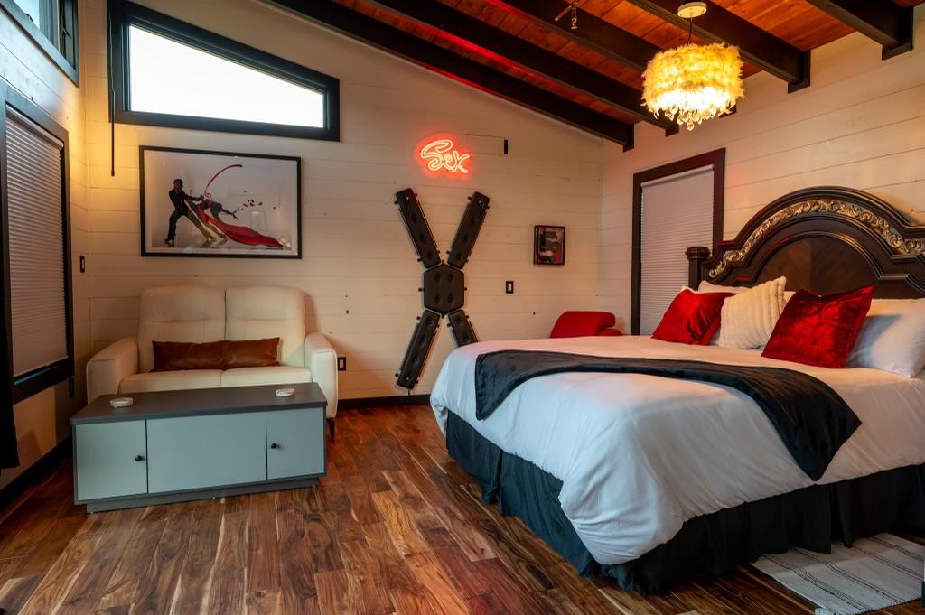 A well-appointed room features a king-size bed adorned with rich red and white bedding. A white leather loveseat is positioned beside a modern coffee table. Large windows allow natural light to fill the space, highlighting the wooden ceiling beams and unique wall decor.