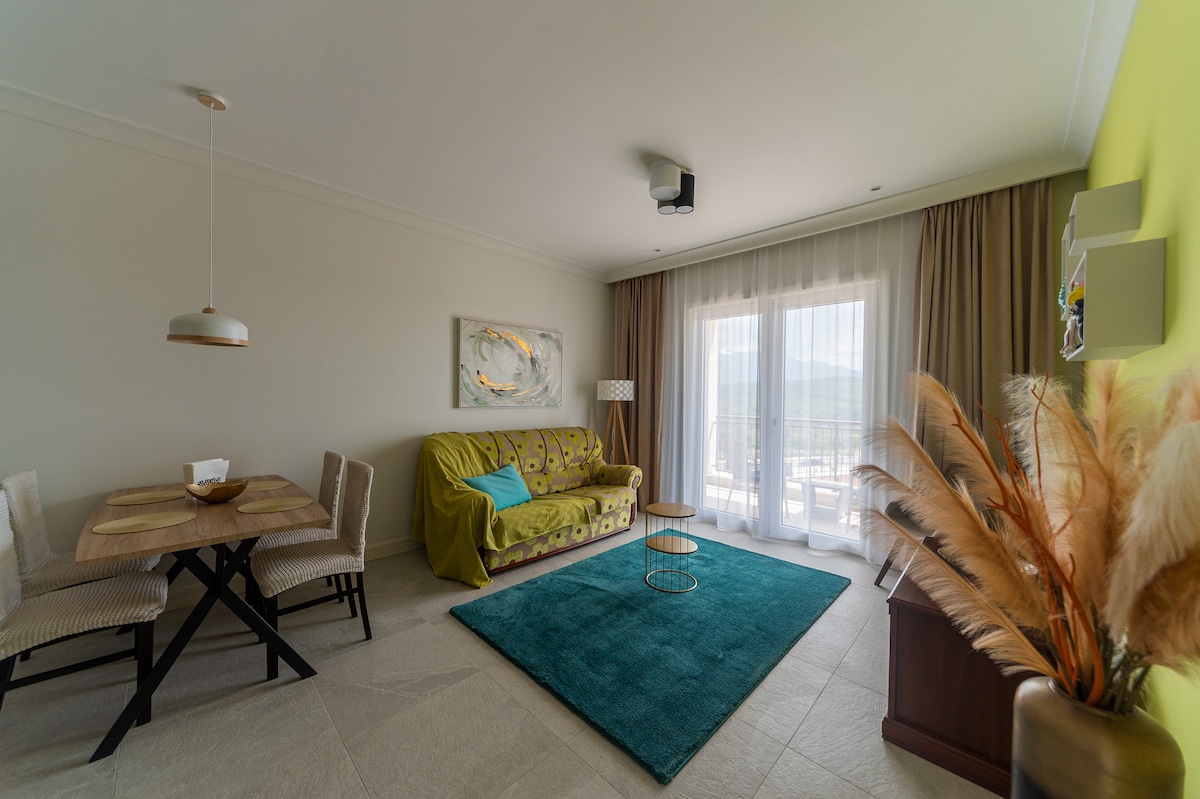 A modern living area features a green sofa and a circular coffee table set on a turquoise rug. A dining table with four chairs is positioned nearby. Large glass doors open to a view outside, while curtains frame the space, adding a soft touch.
