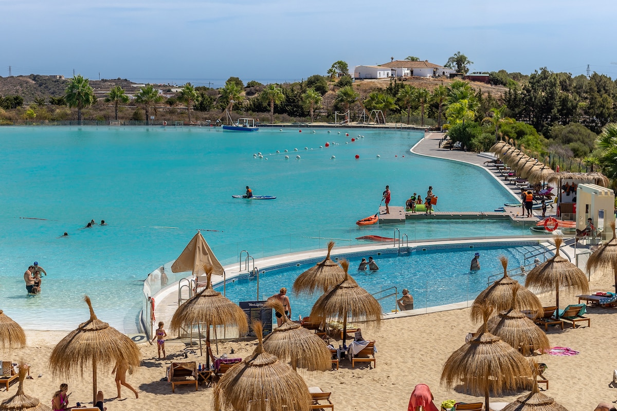 A vibrant artificial lagoon is framed by sandy shores and lush greenery. Lounge chairs with thatched umbrellas provide shaded spots for relaxation. Kayaks and paddleboards are visible on the calm water, while a small swimming area is surrounded by a gentle slope leading to the beach.
