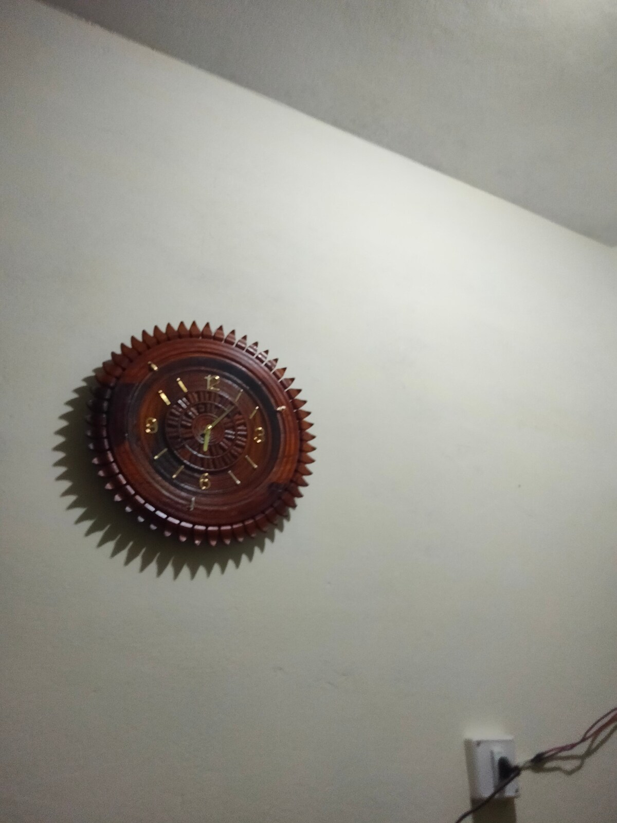 A wooden wall clock is displayed against a light-colored wall. The clock features ornate detailing with golden numbers and hands, providing an elegant touch to the space. An electrical outlet is visible nearby, adding to the functional aspects of the room.
