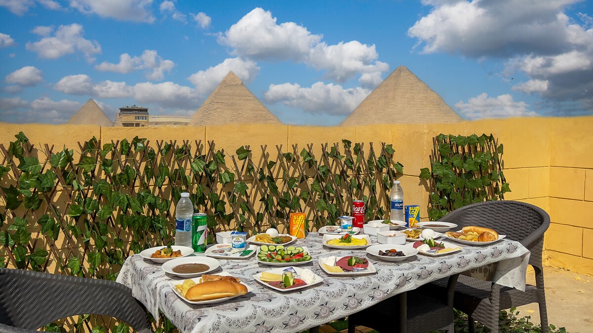 A dining table is set outdoors, covered with various dishes and drinks. Lush greenery rises along the side, while the pyramids of Giza are visible in the background under a clear blue sky with scattered clouds.