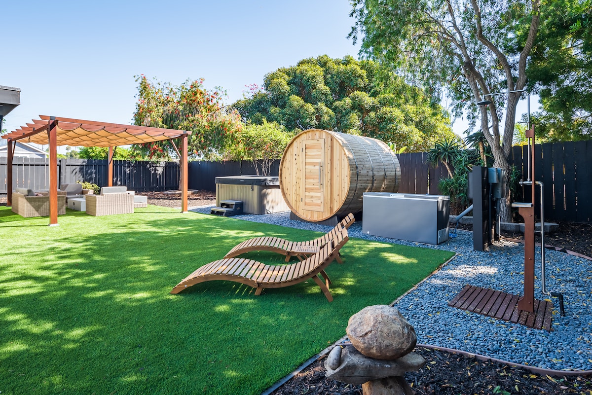 An expansive, secluded backyard features a lush green lawn surrounded by mature trees. A wooden sauna and hot tub are set alongside a sheltered seating area with a canopy. Two lounge chairs provide additional relaxation space near pebbled walkways.