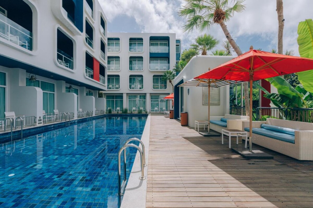 The outdoor pool area is highlighted by a vibrant blue pool, surrounded by a wooden deck. Lounge chairs are arranged along the edge, complemented by shaded seating areas featuring red umbrellas. Lush greenery can be seen in the background, providing a relaxed ambiance.