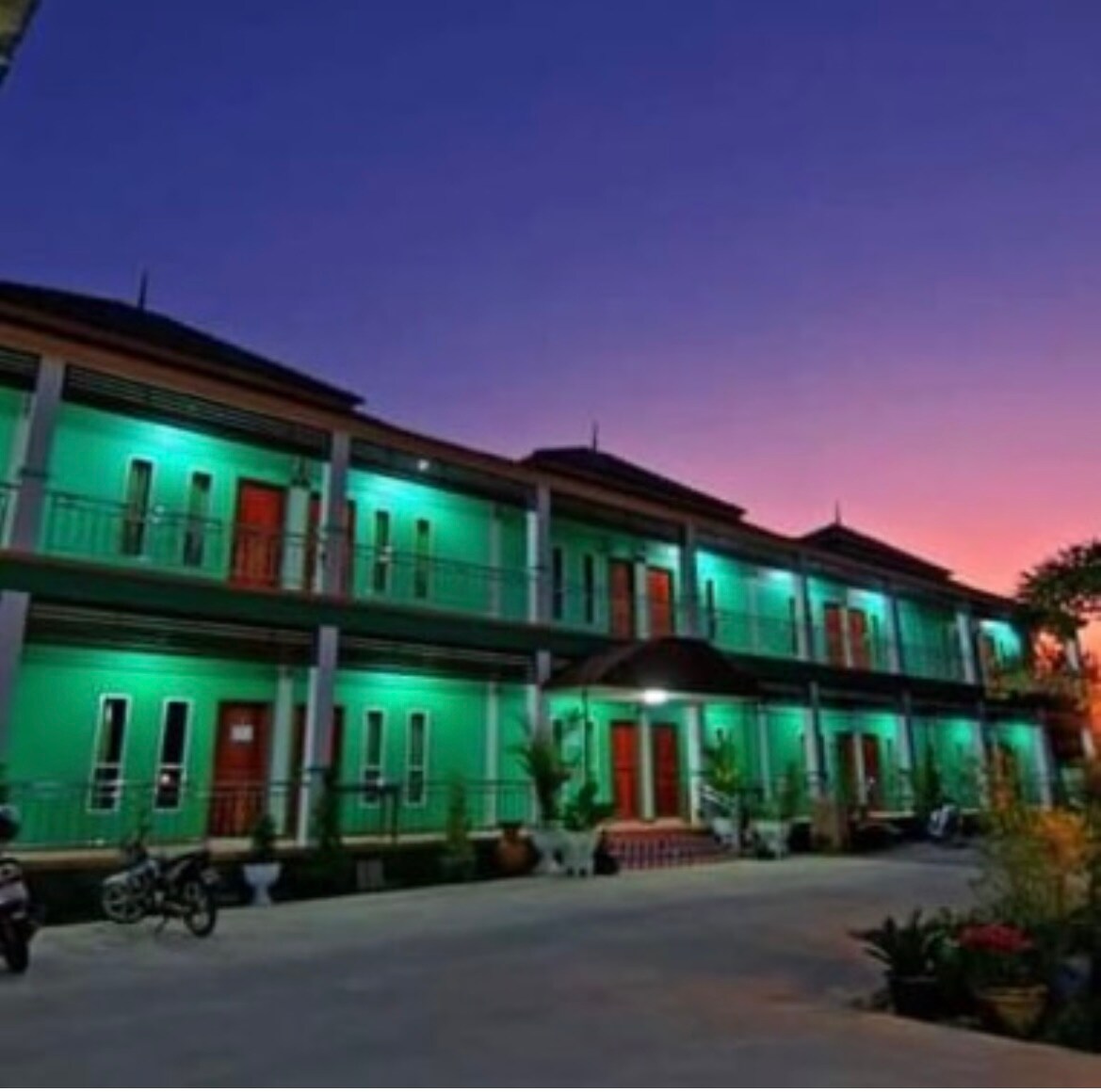 A contemporary two-story building is featured, illuminated with soft green lights against a twilight sky. Balconies are visible on both levels, with multiple doors leading into guest rooms. The surrounding area includes potted plants and parked bicycles, suggesting a welcoming environment.