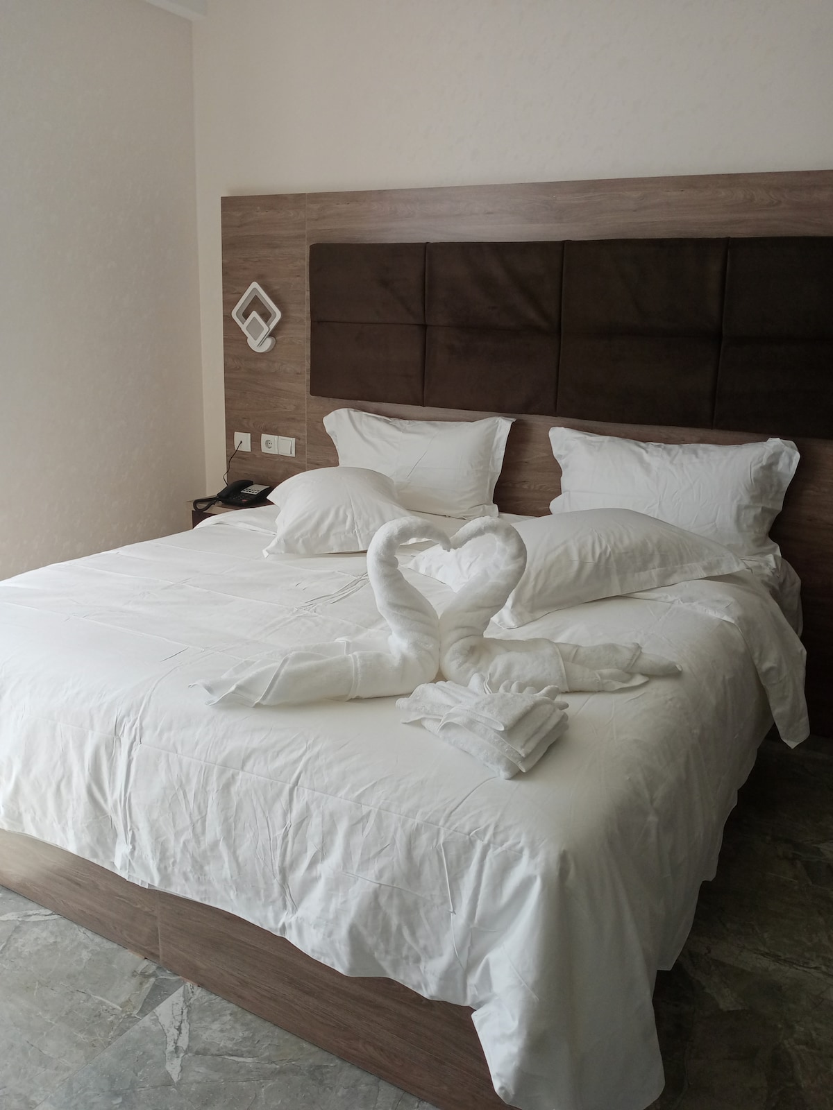 A spacious bed is covered with white linens, featuring decorative towel swans arranged at the center. Pillows are placed neatly at the headboard, which is designed with a warm brown wood finish. The room includes soft lighting and a simple yet elegant decor.