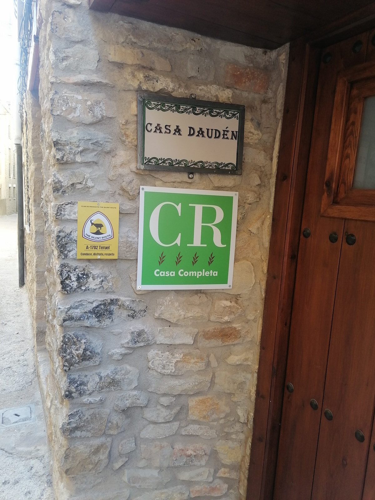 The entrance to Casa Daudén is marked by a rustic stone facade, featuring a wooden door and a sign displaying the property name. A bright green sign indicates 'Casa Completa', while a yellow safety notice is affixed to the wall, contributing to the welcoming ambiance.