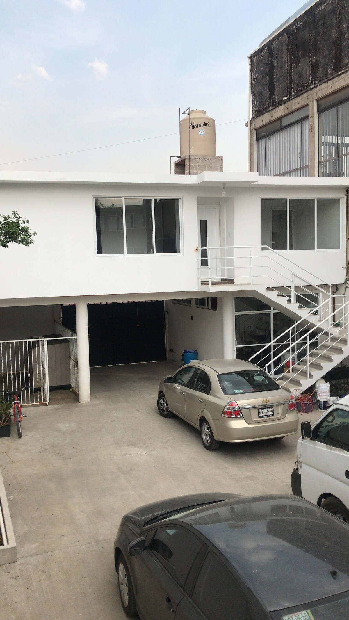 An exterior view of a two-story building is shown, featuring a white façade and a staircase leading to the upper level. A spacious driveway accommodates multiple vehicles, highlighting a designated parking area. Large windows allow natural light to enter the interior spaces.