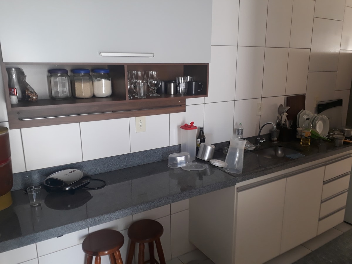 A well-equipped kitchen features a long countertop with various utensils and appliances organized efficiently. Glass jars and containers are neatly arranged on the upper shelf. Two wooden stools provide seating, complementing the modern cabinetry and tiled walls.