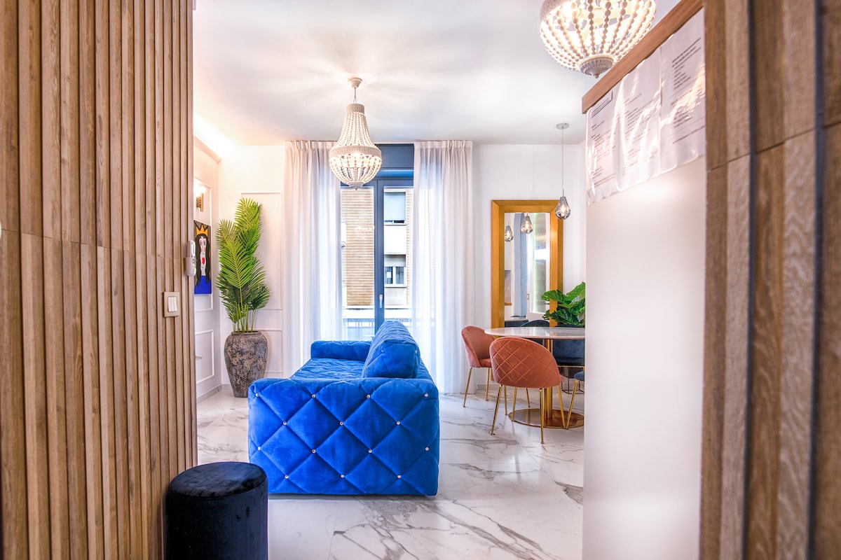 A spacious living area is characterized by a plush blue sofa complemented by elegant lighting fixtures. Large windows invite natural light, while the decor features wooden accents. A dining table with colorful chairs sits at the opposite end, reflecting a modern design aesthetic.