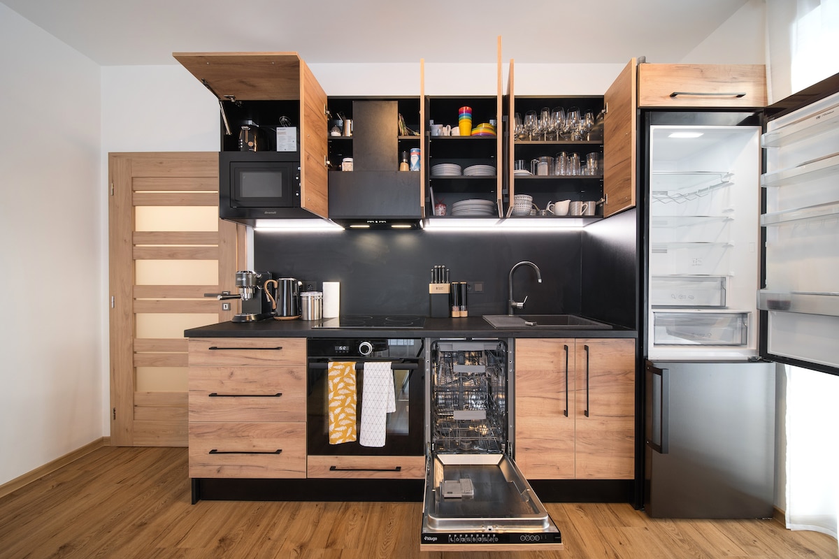 A modern kitchen setup is showcased with wooden cabinets and black countertops. Dishware is neatly arranged on shelves. A dishwasher is shown with its door open, while a refrigerator stands beside it, displaying a clear door and organized compartments. The overall space appears well-lit and functional.