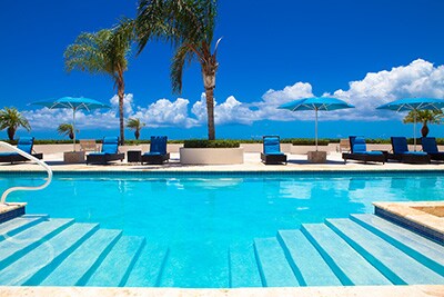 A sparkling saltwater pool is framed by palm trees and blue lounge chairs, inviting relaxation under clear skies. Umbrellas provide shade beside the pool's gentle steps, leading into refreshing water. The backdrop features coastal views and puffy white clouds.