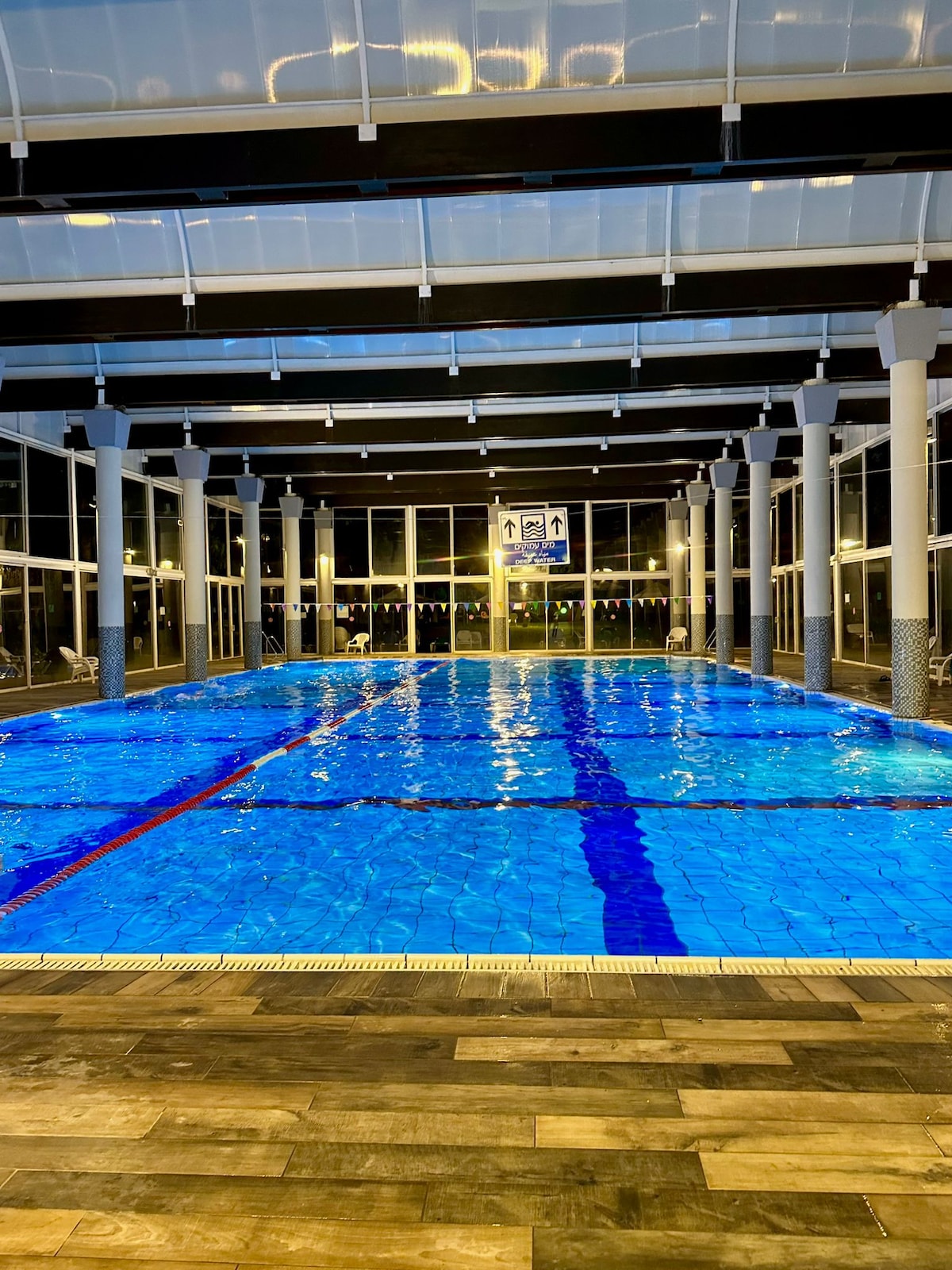 An indoor swimming pool is illuminated with bright blue water, reflecting light from surrounding windows. Lounge chairs are positioned along the edges, and clear signage can be seen on the far wall. A warm ambiance is created by the lighting and glass walls.