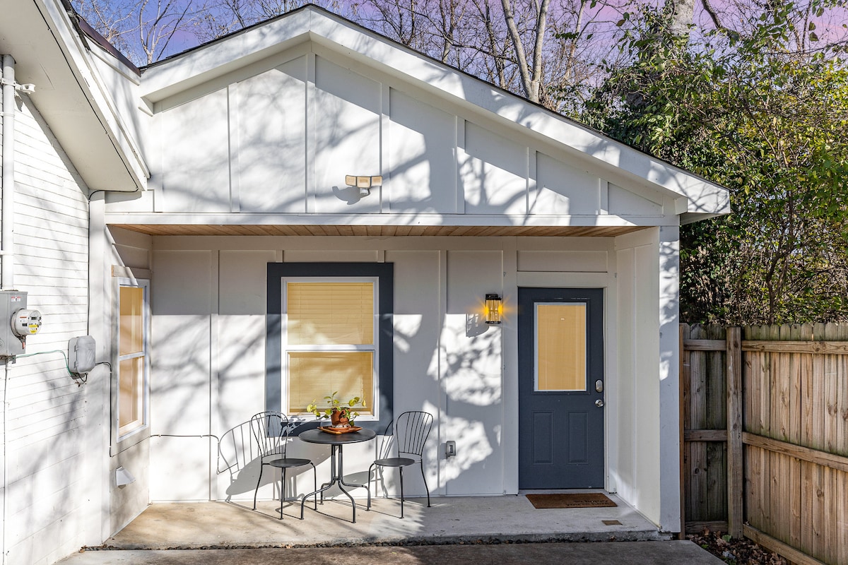 An inviting outdoor area is shown, featuring a small table with two chairs positioned near the entrance. Natural sunlight filters through nearby trees, casting gentle shadows on the white exterior walls. A decorative lamp illuminates the entryway, emphasizing the welcoming atmosphere of the space.