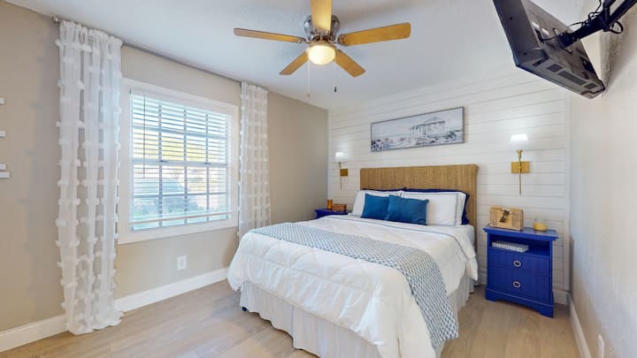 The master bedroom is cozy and peaceful with gauzy curtains, shiplap wall, jute headboard, bright blue nightstands, and soft side lights.