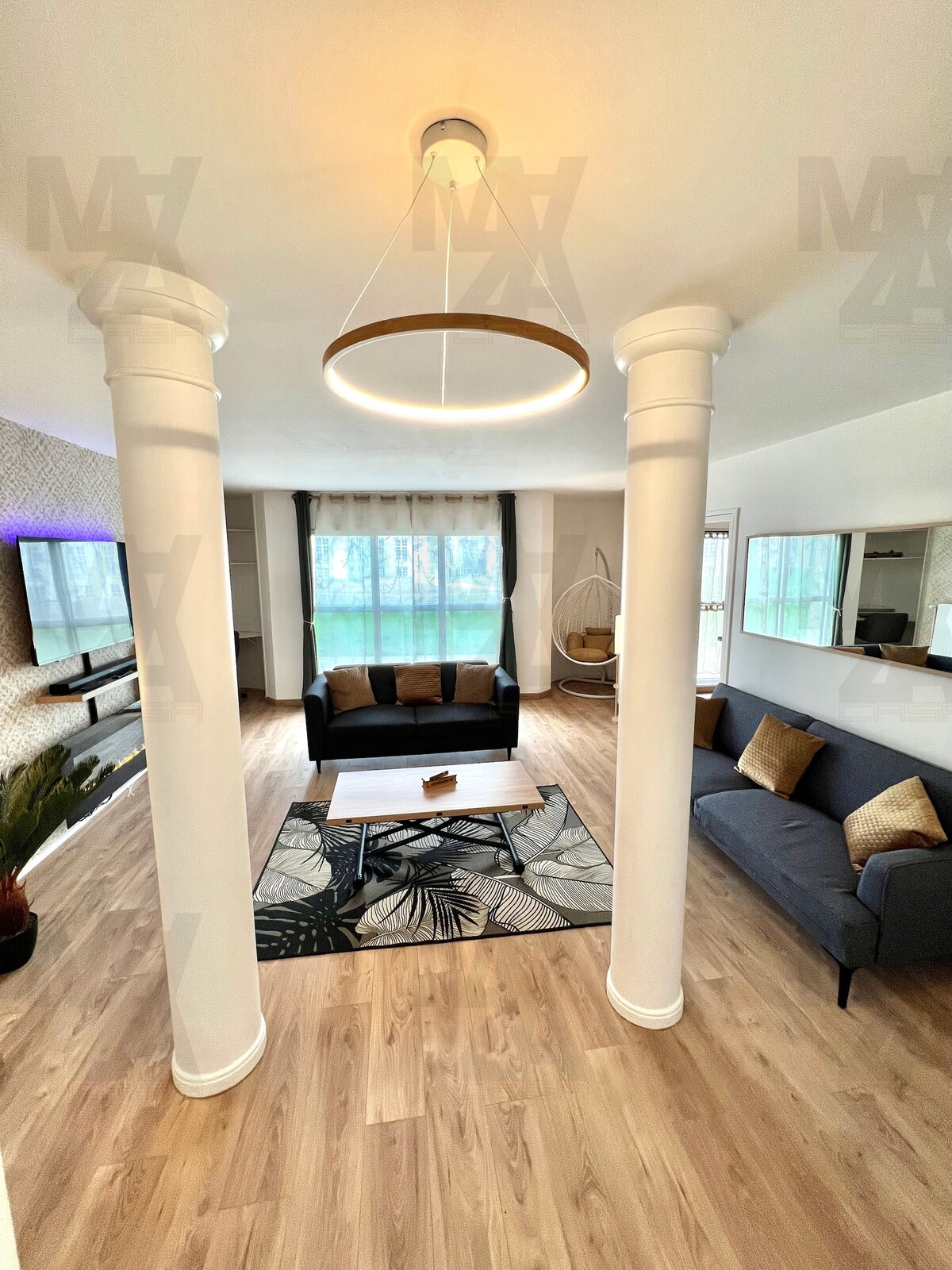 The living area features two distinct sofas, a central table, and an artistic rug. Tall windows allow natural light to illuminate the space, complemented by decorative wall elements. A unique hanging chair adds a cozy touch to the room's layout.