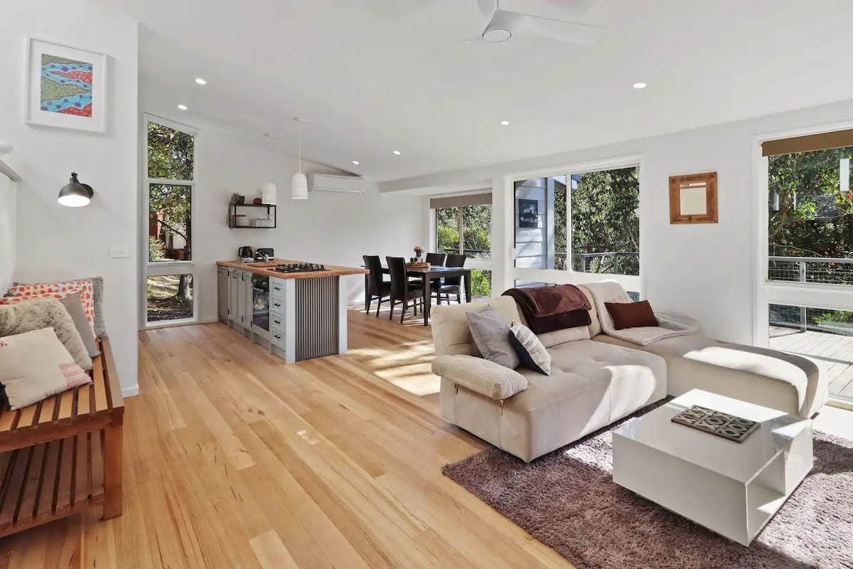 An open-plan living space features a neutral-colored sectional sofa and a coffee table, complemented by natural wood flooring. Large windows provide ample light and views of the surrounding greenery. A dining area with a wooden table is visible in the background.