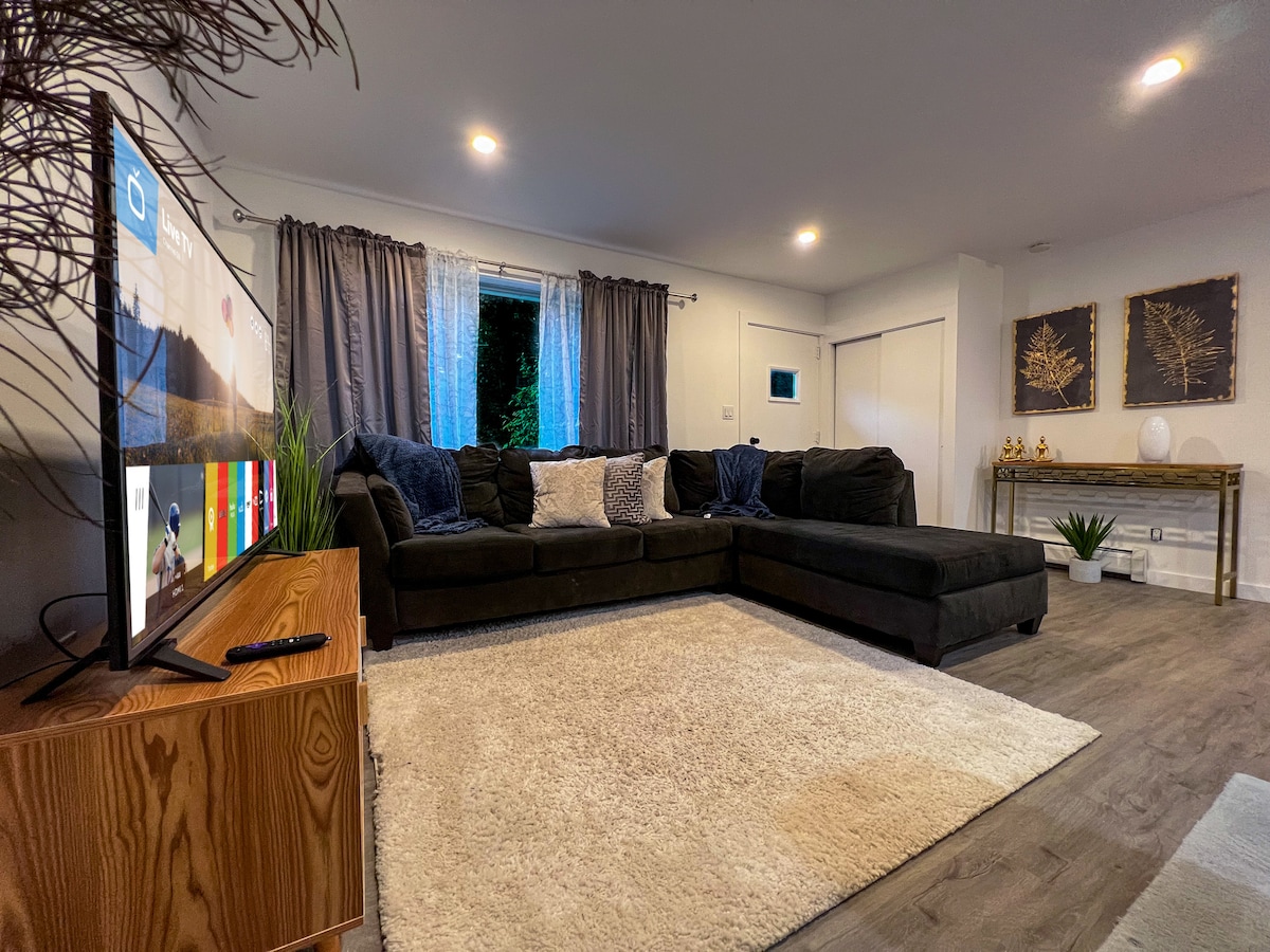 A spacious living room features a large sectional couch positioned near a modern TV. Soft lighting illuminates the area, while curtains frame the window. A plush area rug covers the floor, complemented by decorative accents on the walls.